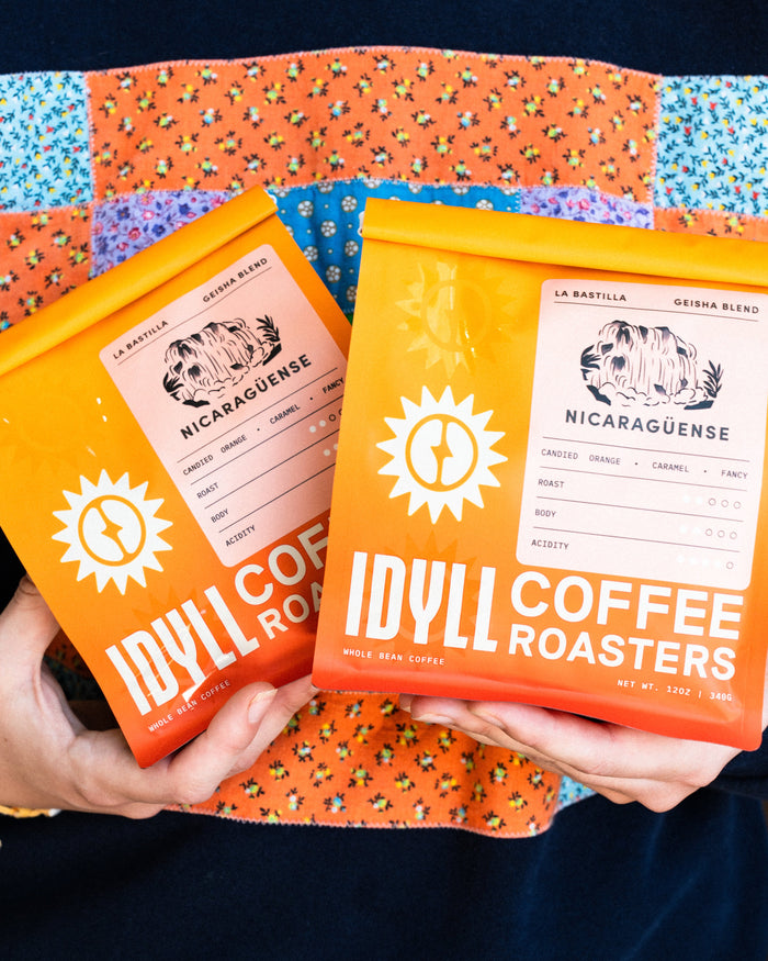Nicaragüense La Bastilla Geisha Blend coffee bags in bright orange packaging held against a colorful patchwork cloth