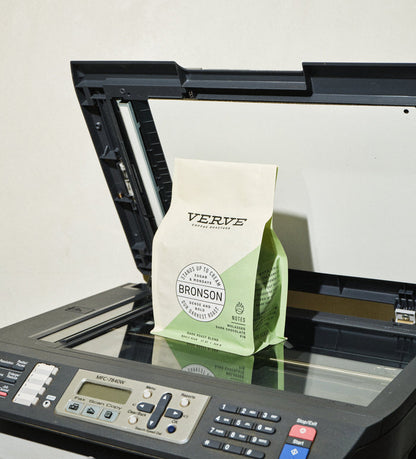 Bronson coffee bag on a marble counter with coffee beans spilled and a copier in the background
