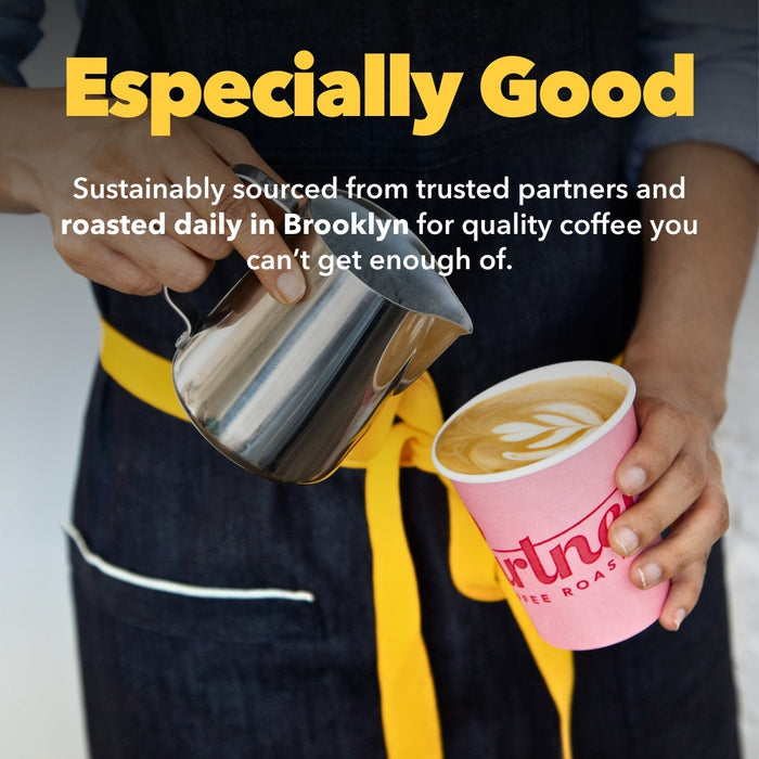 Barista pours milk from a stainless pitcher into a pink coffee cup with latte art, wearing a dark apron with a yellow belt.